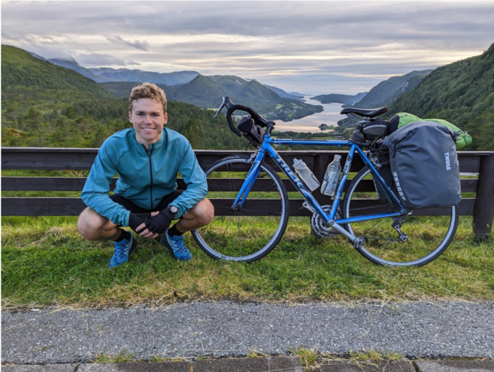young man posing next to a bike with mountains and lake landscape at the back