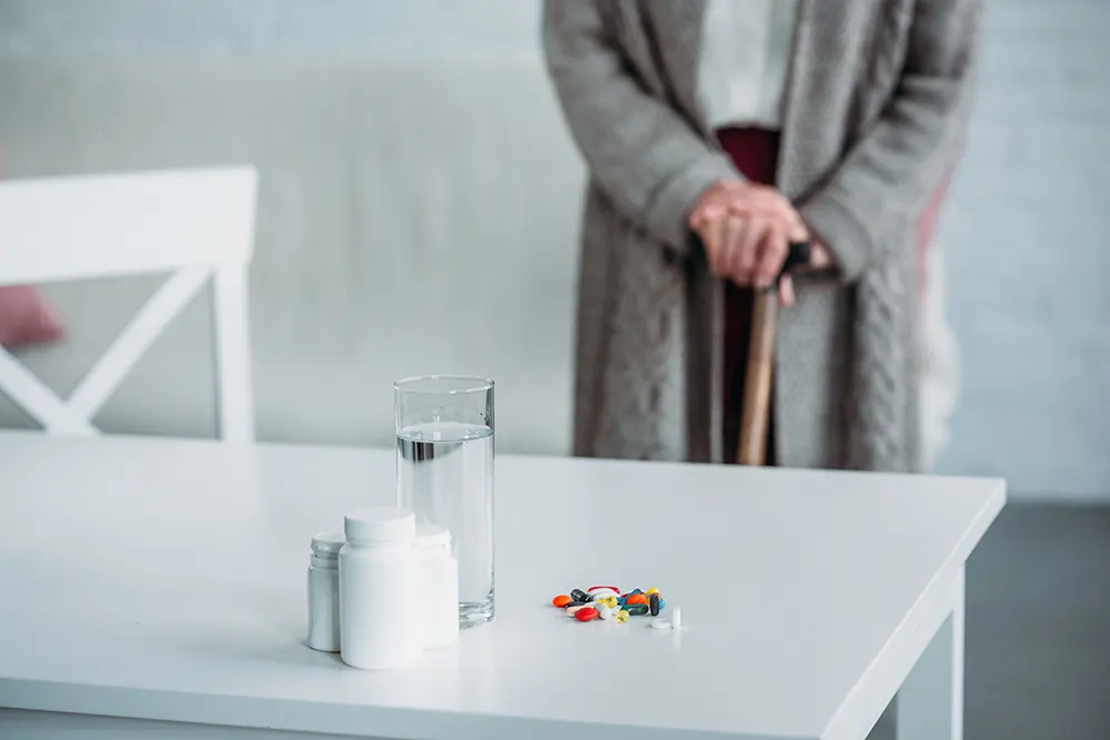Medisinar oppstilt på ei bordplate. I bakgrunnen står ei eldre kvinne med stokk