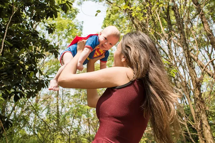 Kvinne som lyftar ein baby i veret utandørs. babyen har på seg Supermann-drakt