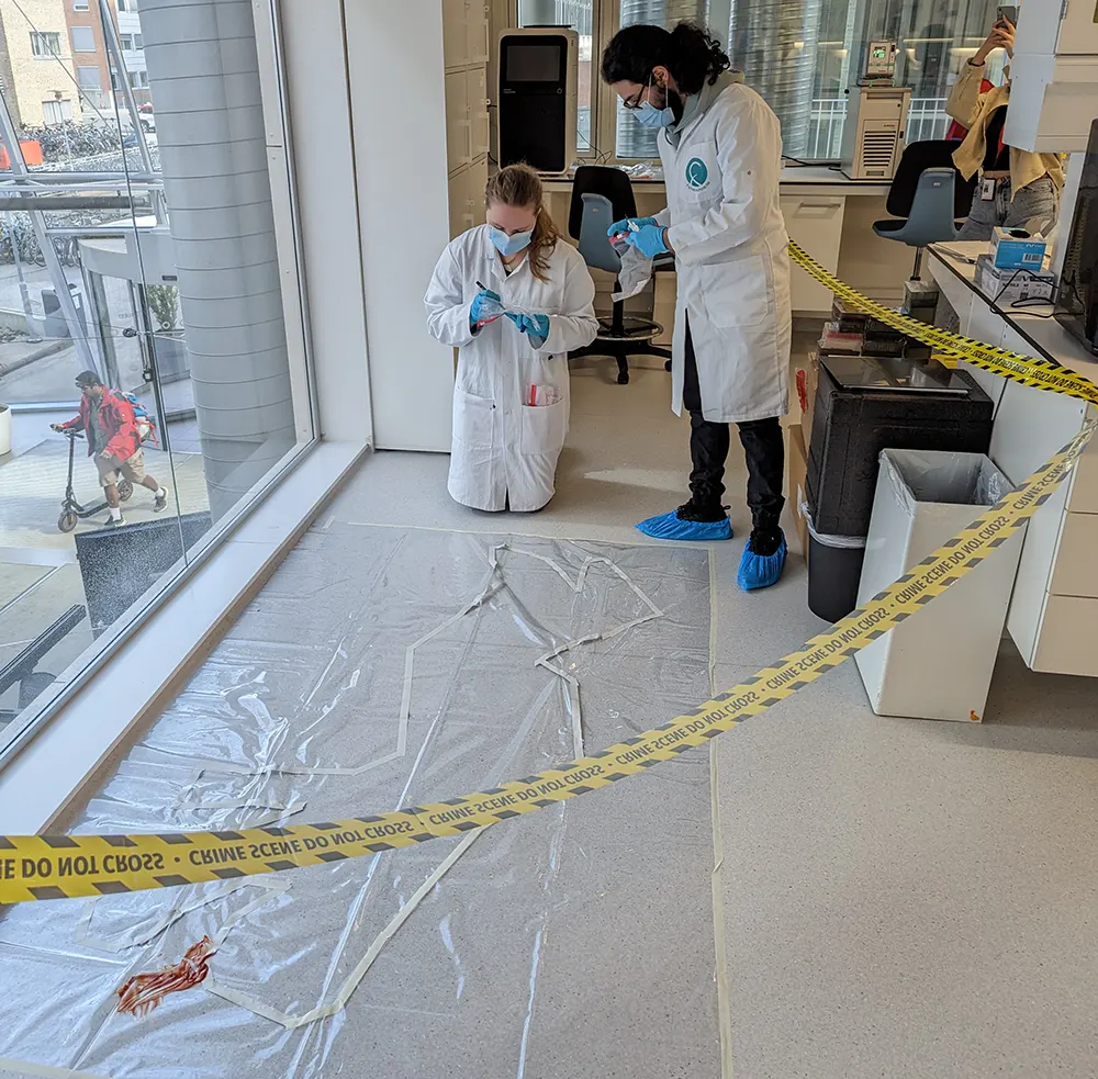 To studentar i labfrakk bøygd over eit omriss av ein menneskekropp på golvet