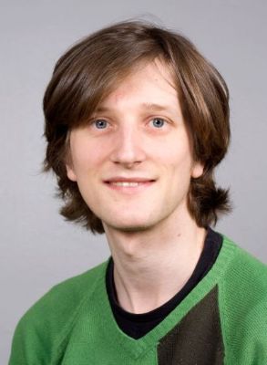 A portrait of a man with brown hair, wearing a green sweater and smiling.