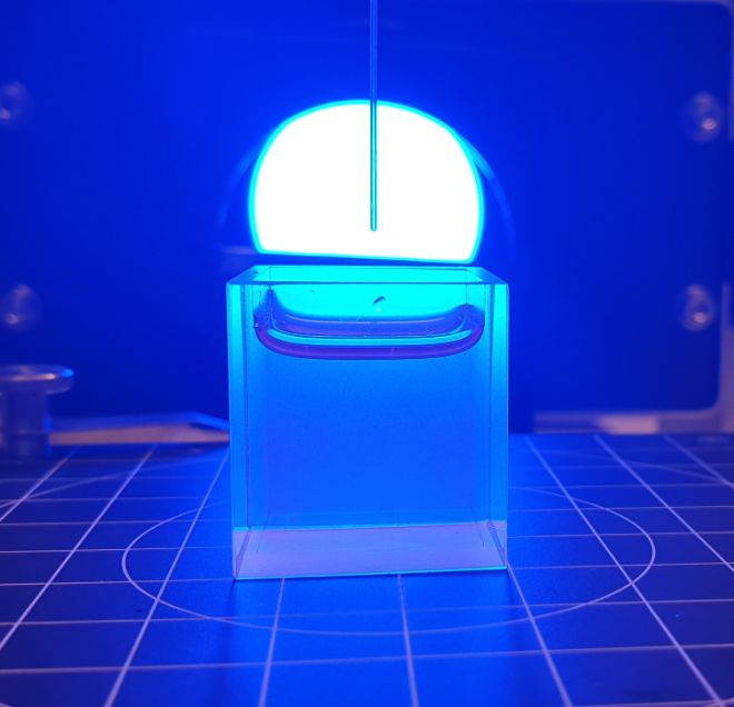 A see-through box with water in it with a thin pipe hanging vertically above. A blue light is coming from a lamp behind the box.